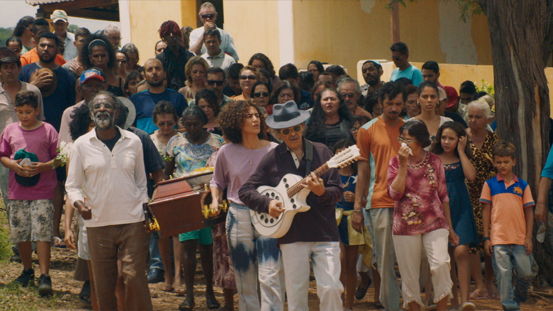 Trailer still frame from Bacurau, people walking behind man playing guitar